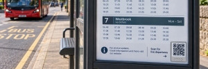 a bus stop timetable on a bus shelter with a QR code with a bus in the distance.